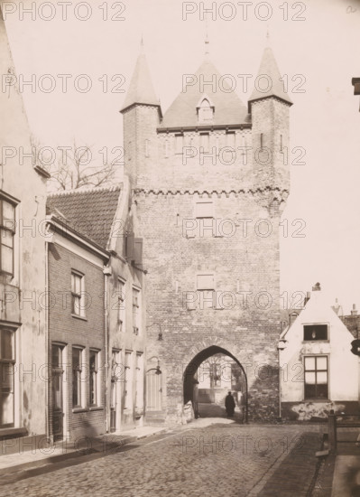 View of the Dijkpoort in Hattem, photograph, annotation: 'a, Hattem', annotation: 'Hattum, Dijkspoort in restored condition Feb. 1910', Hattem, 1910, photographer, Attributed to, paper, pasteboard, daylight collodion silver print, height 218 mm x width 159 mm, city-gate (city(-scape) with figures, staffage), Dijkpoort, A tall brick gatehouse with two pointed turrets and a steep central roof rises above a cobbled street, showing small shuttered windows and an arched passage at its base; flanking the gatehouse are rows of narrow brick and plaster buildings with tiled roofs, rectangular windows, doors, and wall-mounted lamps; a solitary figure stands in the shadowed archway, and leafless tree branches are visible behind the buildings.