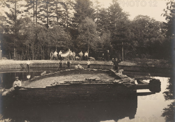 Foundation of a hunting lodge in Heelsum, photograph, annotation: 'F', annotation: 'Heelsum, Foundation of a hunting lodge? found at the Duno country estate', Heelsum, 1890 - 1920, photographer, Attributed to, paper, pasteboard, gelatin silver print, developed gelatin silver print, height 158 mm x width 219 mm, ruin of a dwelling, house, castle, landscape with ruins (city(-scape) with figures, staffage), A wide sepia photograph shows a partially built brick structure extending into calm water with reflections of the structure and surrounding trees; men in period clothing are positioned around the site, some standing on the far bank near a row of trees, others standing or sitting on the brickwork and at the water’s edge, one man seated on a corner of the brickwork holding an object, another man standing on the bank holding long poles, and a small group near the tree line holding a large draped fabric; wheelbarrows and tools are visible behind the group on the bank, the background is fil...
