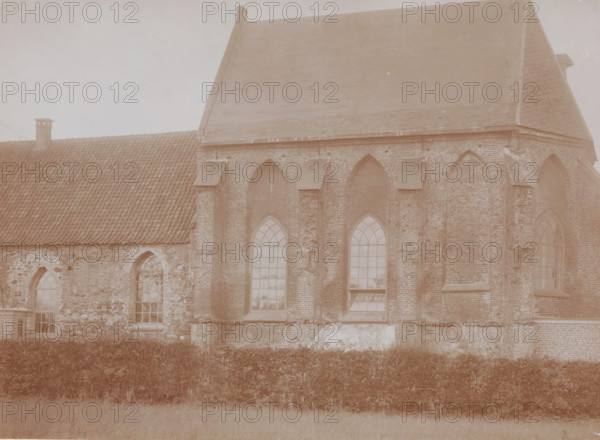 View of the choir of the Reformed Church in Andelst, photograph, annotation: 'Church of the Dutch Reformed congregation in Andelst, Gelderland, Nov 1910, Municipality of Valburg', Andelst, 1910, photographer, Attributed to, paper, pasteboard, daylight collodion silver print, height 158 mm x width 217 mm, parts of church exterior and annexes: choir, A sepia-toned photograph of a large brick building with steep tiled roofs and arched windows, showing textured brickwork and stone patches on the lower wall, a row of tall pointed-arch windows with multi-pane glass, a smaller tiled roof section with a chimney, and a hedge running along the base of the building with a grassy area in the foreground.
