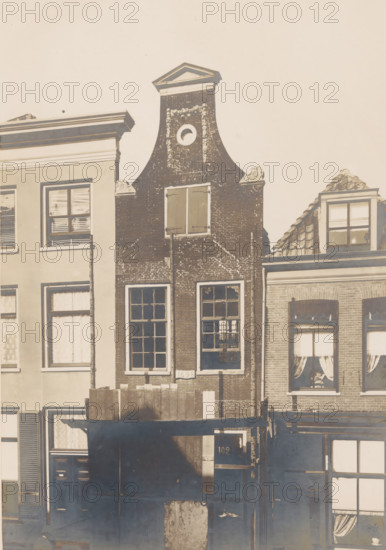 Façade of Langestraat 102 in Amersfoort, photograph, annotation: 'Amersfoort (U.), Langstraat No 102, Oct 1902, Aug 1902', Amersfoort, 1902, photographer, Attributed to, paper, pasteboard, gelatin silver print, height 217 mm x width 148 mm, urban housing, façade (of house or building), A narrow brick building with a curved stepped gable and a triangular pediment at the top stands between adjacent buildings, featuring a small circular opening near the top and decorative stone scrolls on either side of the gable, tall multi-pane windows on the lower floors with shutters on the upper window, and visible brick texture and weathering; neighboring facades include a lighter smooth-stucco building with rectangular windows and blinds on the left and a building with tiled roof dormer and windows with curtains on the right, while wooden boards and a dark awning or canopy cover part of the lower frontage.