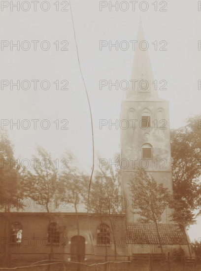 View of the Reformed Church in Schalkwijk, photograph, annotation: 'Dutch Reformed Church in Schalkwijk, Aug 1911, (U.)', Schalkwijk, 1911, photographer, Attributed to, paper, pasteboard, daylight collodion silver print, height 213 mm x width 157 mm, height 326 mm x width 234 mm, parts of church exterior and annexes: tower, A tall building with a steep pointed roof and a clock face on its upper section rises above a lower structure with arched windows and a tiled roof; the taller section has arched window openings and a louvered vent, and the facade shows patches of discoloration; trees with blurred foliage stand in front of the buildings, and a thin vertical cable or wire runs down the left side of the image; the overall scene appears washed in a muted, sepia-toned palette.