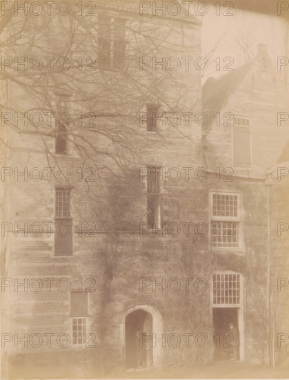 Part of IJsselstein Castle, photograph, annotation: 'IJsselstein', annotation: 'Castle in IJsselstein, demolished 1888', IJsselstein, c. 1888, photographer, Attributed to, paper, pasteboard, albumen print, height 233 mm x width 178 mm, ruin of a dwelling, house, castle, landscape with ruins (city(-scape) with figures, staffage), IJsselstein Castle, A faded photograph of a brick building facade with multiple windows and doorways, bare tree branches extending across the upper portion, a rounded arch doorway near the lower center with a person standing inside the shadowed opening, a taller rectangular opening beside it with another person partly visible in darkness, several rectangular windows with grid-like panes and some shuttered openings, a gabled roof section on the right, and climbing vines or vegetation covering parts of the lower and middle walls.