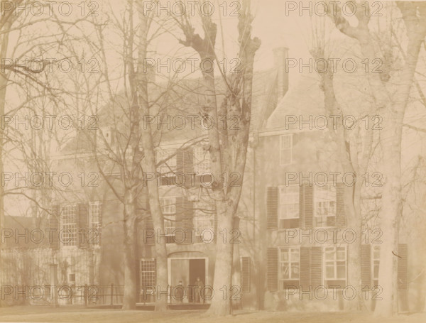 View of IJsselstein Castle, photograph, annotation: 'Castle in IJsselstein, demolished 1888', annotation: 'IJsselstein', IJsselstein, c. 1888, photographer, Attributed to, paper, pasteboard, albumen print, height 174 mm x width 230 mm, ruin of a dwelling, house, castle, landscape with ruins (city(-scape) with figures, staffage), IJsselstein Castle, A faded photograph of a large building partially obscured by many leafless trees, showing a facade with shuttered windows, a pitched roof with a chimney, and a doorway with a small porch; two standing figures are visible on the porch beneath the trees; a streetlamp is positioned near the center, and a low fence runs along the front of the building.