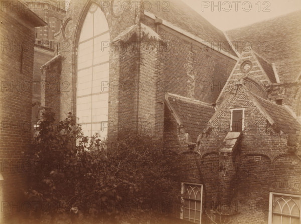 South side of the Old St. Nicholas Church in IJsselstein, photograph, annotation: 'Church in IJsselstein, South side, tower, transept, consistory, Nov. 1890', annotation: 'IJsselstein', IJsselstein, 1890, photographer, Attributed to, paper, pasteboard, albumen print, height 177 mm x width 233 mm, church (exterior), Oude Sint-Nicolaaskerk, A sepia-toned view of an old brick building complex showing a large arched window with a grid of panes set in a tall wall, adjacent buttressed brickwork with weathered and mossy patches, steep tiled roofs, a small gabled section with a dark shuttered window framed by lighter trim, decorative brick patterns on a triangular gable, arched openings beneath a row of roof tiles, two arched glazed windows lower on the wall, and dense foliage and branches growing in front of the lower portion of the walls.