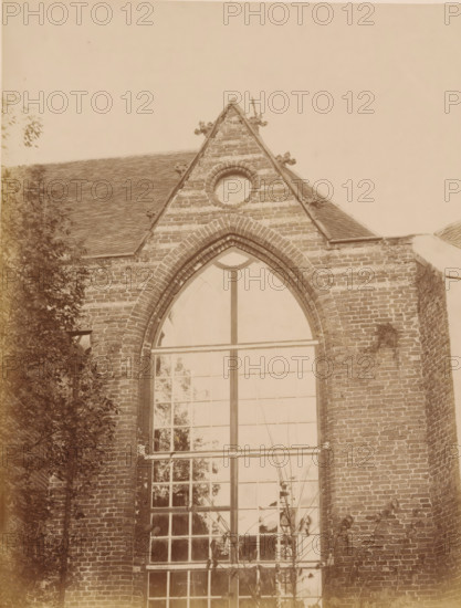 Part of the choir of the Old St Nicholas Church in IJsselstein, photograph, annotation: 'Church in IJsselstein, Unplastered ... of the choir, Nov. 1890', annotation: 'IJsselstein', IJsselstein, 1890, photographer, Attributed to, paper, pasteboard, albumen print, albumendruk, height 230 mm x width 176 mm, parts of church exterior and annexes: choir, Oude Sint-Nicolaaskerk, A section of a brick building with a steep gabled roof and decorative stone accents under the gable peak, a circular masonry feature near the top of the gable, and a tall arched window with multiple panes occupying the central facade; portions of climbing plants and tree branches frame the left side and base of the wall, and the brickwork shows weathering and mortar lines.