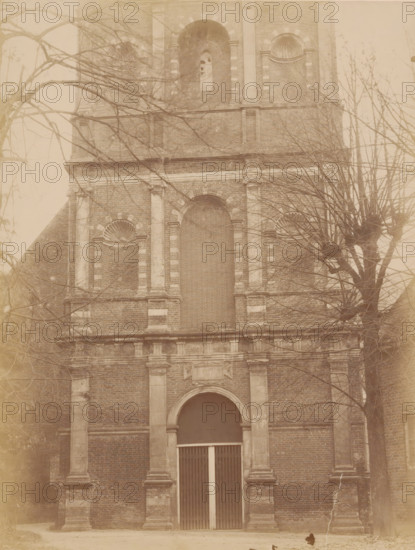 Tower of the Old St. Nicholas Church in IJsselstein, photograph, annotation: 'IJsselstein', annotation: 'Tower in IJsselstein, Westfront (lower part), Nov. 1890', IJsselstein, 1890, photographer, Attributed to, paper, pasteboard, albumen print, height 232 mm x width 176 mm, parts of church exterior and annexes: tower, Oude Sint-Nicolaaskerk, A tall brick building facade with stone columns and decorative arches, leafless tree branches partially obscuring the view, a large arched doorway with vertical slatted doors, recessed niches with shell-shaped reliefs, rectangular stone detailing around windows and along the facade, and bare ground in front of the entrance.