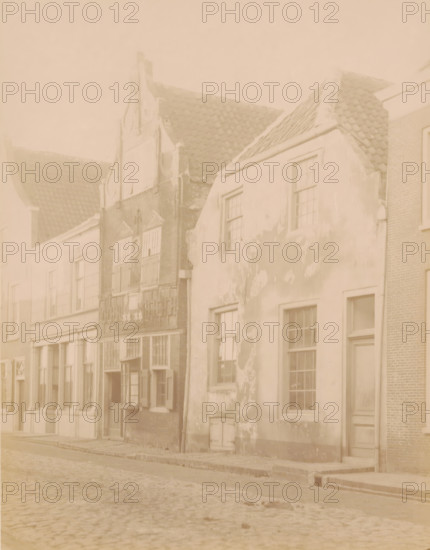 Houses in the Benschopperstraat in IJsselstein, photograph, annotation: 'IJsselstein', annotation: 'House on Benschopperstraat in IJsselstein demolished 1889', IJsselstein, c. 1889, photographer, Attributed to, paper, pasteboard, albumen print, height 231 mm x width 179 mm, urban housing, street, A row of old buildings lines a cobbled street, showing varied facades and rooflines with tiled roofing; one building has a stepped gable and decorative brickwork, another has a plastered exterior with patches of peeling paint and several multi-pane windows, and a narrow doorway set into its wall; wooden window shutters appear open on one facade, while stone and brick details are visible around entrances and lower walls; the street foreground shows uneven cobblestones and a raised pavement edge.