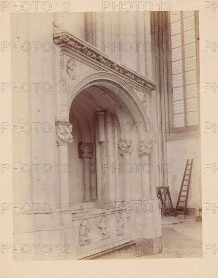 View of the Holy Sepulchre in the Cathedral of Utrecht, photograph, annotation: 'Utrecht', annotation: 'Cathedral in Utrecht, Sept 1891, The Holy Sepulchre', Cathedral of Utrecht, 1891, photographer, Utrecht, Attributed to, paper, pasteboard, albumen print, height 232 mm x width 175 mm, interior of church, grave-building, monumental tomb, A stone archway with carved decorative molding and sculpted figures sits against a wall of tall vertical stone columns, a section of a carved stone panel below the arch shows recessed relief figures, carved column capitals flank the arch, and a tall leaded window with diamond panes appears to the right; a wooden ladder leans against the wall near a small folding workstand on the floor.