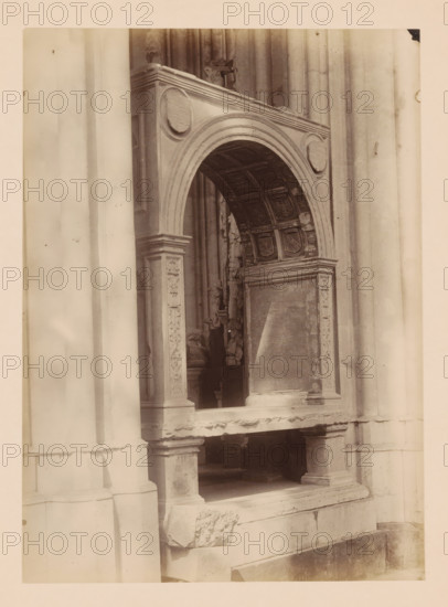 Grave monument in the Dom of Utrecht, photograph, annotation: 'Utrecht', annotation: 'Cathedral in Utrecht, grave monument, Sept 1891', Dom of Utrecht, 1891, photographer, Utrecht, Attributed to, paper, pasteboard, albumen print, height 239 mm x width 173 mm, interior of church, grave-building, monumental tomb, A sepia-toned photograph of a stone architectural feature showing a carved archway with decorative panels and reliefs; the arch has an ornate coffered underside with square recessed panels and shield-like motifs, and circular medallions set into the outer corners; fluted columns and pilasters with carved floral and geometric ornament flank the opening, and a recessed rectangular panel with worn surface sits beneath the arch; the stone shows weathering and surface irregularities, and a ledge and base with visible joints and chipped edges occupy the lower portion of the image.