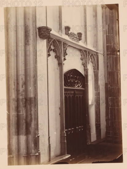 Portal in the Cathedral of Utrecht, photograph, annotation: 'Cathedral in Utrecht, Sept 1891', annotation: 'Utrecht', Cathedral of Utrecht, 1891, photographer, Utrecht, Attributed to, paper, pasteboard, albumen print, height 238 mm x width 173 mm, interior of church, A vertical sepia photograph of an architectural interior showing a section of a stone wall with tall fluted columns on the left and right, ornate carved capitals and arch moldings, a pointed arch doorway with decorative wrought elements in its upper panel, slender clustered shafts and carved foliage motifs along the archway, a ledge running above the doorway with sculpted corbels and small carved heads, a row of wooden seating with carved ends visible in the lower right, and areas of light and shadow across the stone surfaces.