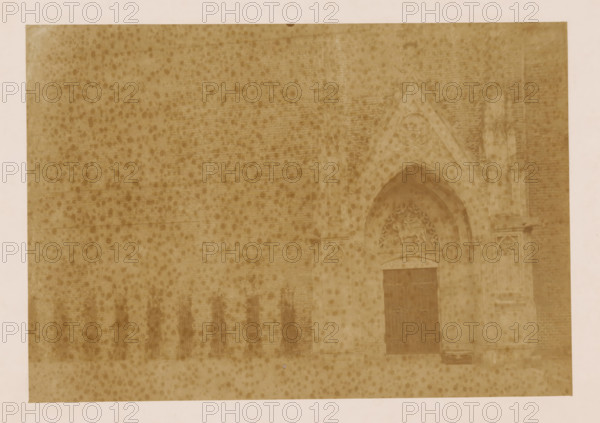 West wall of the cloister at the Dom of Utrecht, photograph, annotation: 'Utrecht', annotation: 'West wall of the cloister in Utrecht', Dom of Utrecht, 1890 - 1910, photographer, Utrecht, Attributed to, paper, pasteboard, albumen print, height 161 mm x width 232 mm, cloisters, monastery, A faded sepia photograph showing a textured brick wall with darker spotting across its surface and a pointed stone archway set into the wall on the right side, the archway featuring carved decorative stonework and a recessed doorway with a dark wooden door, flanking stone columns and sculpted relief above the arch, with part of a pale border visible around the image.