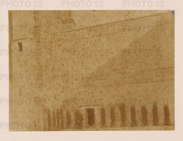 West wall of the cloister at the Dom of Utrecht, photograph, annotation: 'West wall of the cloister in Utrecht, Dec 1900', annotation: 'Utrecht', Dom of Utrecht, 1900, photographer, Utrecht, Attributed to, paper, pasteboard, albumen print, height 160 mm x width 220 mm, cloisters, monastery, A sepia-toned photograph of a large brick wall with faint horizontal lines and a small doorway near the bottom center, flanked by a row of tall, bundled vertical objects against the wall, with a person standing near the doorway wearing a long dark coat and a hat; the surface of the image shows numerous dark speckled spots and a lighter border around the edges.