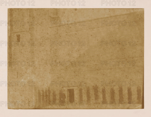 West wall of the cloister at the Dom of Utrecht, photograph, annotation: 'West wall of the cloister in Utrecht (Utrecht), Dec 1900', annotation: 'Utrecht', Dom of Utrecht, 1900, photographer, Utrecht, Attributed to, paper, pasteboard, albumen print, height 170 mm x width 230 mm, cloisters, monastery, A faded sepia-toned rectangular photograph with a wide lighter border shows a large brick wall and a sloped roof occupying most of the frame, the surface marked by scattered round spots and subtle streaks; at the lower center a dark doorway is visible with two faint silhouetted figures standing nearby, and a row of dark vertical bundles or stakes leans against the wall to the right of the doorway.