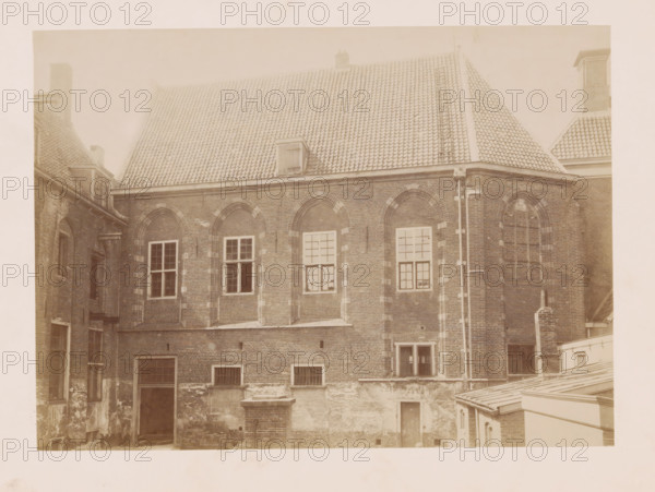 View of the former Agnieten Convent in Utrecht, photograph, annotation: 'Church of the former St. Agnieten Convent, now a military building', annotation: 'Utrecht', Utrecht, 1890 - 1910, photographer, Attributed to, paper, pasteboard, albumen print, height 167 mm x width 230 mm, cloisters, monastery, A sepia-toned photograph of an older brick building showing a large tiled roof with a small dormer and a vertical ladder-like feature, beneath which a row of arched brick window niches contains rectangular windows with multi-pane glazing, some open and some closed; the brick façade shows areas of lighter and darker brickwork and patches of wear, with a vertical downpipe running beside a corner, and lower walls displaying small barred openings and a recessed doorway; adjacent rooftops and chimneys frame the sides, and a portion of a corrugated roof structure appears in the lower right foreground.