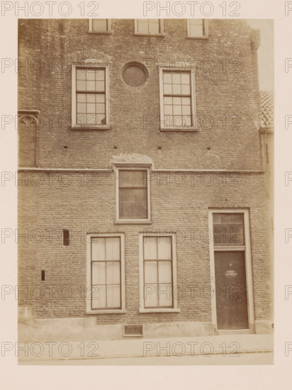 Part of the former Agnieten Convent in Utrecht, photograph, annotation: 'Utrecht', annotation: 'Utrecht, Former St. Agnieten Convent now military building, part bordering a, d church, June 1902', Utrecht, 1902, photographer, Attributed to, paper, pasteboard, albumen print, height 230 mm x width 170 mm, cloisters, monastery, A brick building facade with multiple rectangular windows framed in light trim, a circular recessed opening above two upper windows, a doorway with a darker door set into the brick, narrow vertical vents in the lower brick, a horizontal stone band separating upper and lower sections, a small rectangular grille near the base, and a tiled roof edge visible at the right.
