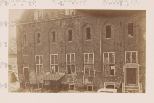 View of the former Agnieten Convent in Utrecht, photograph, annotation: 'Utrecht', annotation: 'Utrecht (U.), Former St. Agnieten Convent now a military building, part bordering a, d church, June 1902', Utrecht, 1902, photographer, Attributed to, paper, pasteboard, albumen print, height 139 mm x width 230 mm, cloisters, monastery, A sepia-toned image of a brick building facade showing multiple vertical window openings with varied panes and some open shutters, a tiled roof with dormer windows at the top, a vertical drainpipe running down the wall, areas of peeling and weathered plaster on the lower brickwork, a small corrugated awning supported by posts above a ground-level opening, scattered wooden beams or planks on the ground, and a few people visible through one of the open lower windows.