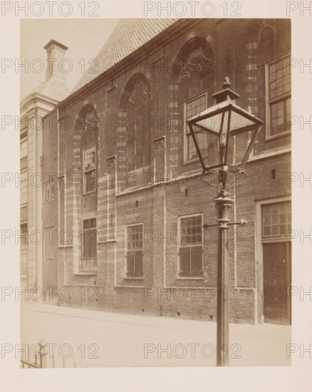 Church of the former St. Agnieten Convent in Utrecht, photograph, annotation: 'Utrecht', annotation: 'Utrecht, Church of the former St. Agnieten Convent, June 1902, now a military building', Utrecht, 1902, photographer, Attributed to, paper, pasteboard, albumen print, height 220 mm x width 170 mm, church (exterior), street lighting, cloisters, monastery, A sepia-toned photograph showing a brick building facade with tall arched windows and rectangular multi-pane windows, decorative stonework around the arches, and a tiled roof visible at the top left; a metal street lamp with a square lantern and glass panes stands in the foreground on a pole with decorative fittings; part of a lighter-colored adjacent building with a chimney and cornice appears at the left edge, and a plain doorway is visible at the right edge; the ground in front of the building is light and smooth, and a short section of a metal railing is visible at the bottom left.