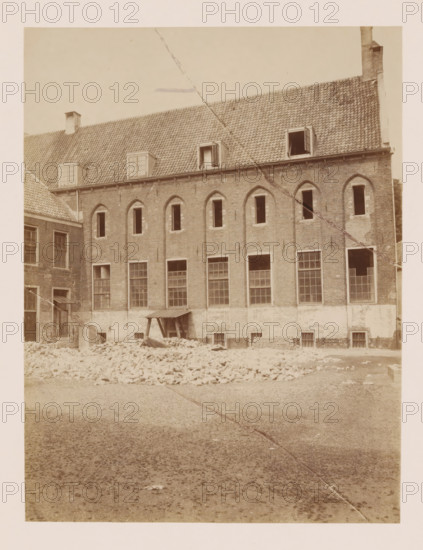 Former St. Agnieten Convent in Utrecht, photograph, annotation: 'Utrecht', annotation: 'Utrecht, Former St. Agnieten Convent now a military building, June 1902', Utrecht, 1902, photographer, Attributed to, paper, pasteboard, albumen print, height 219 mm x width 166 mm, cloisters, monastery, A sepia-toned photograph of the exterior wall of a multi-story brick building with a tiled sloping roof and several dormer windows, some open, and a chimney at one end; the facade shows arched window openings on the upper level and larger rectangular windows on the lower level, some with panes intact and some open or missing; in front of the building there is a pile of loose bricks and rubble, a small wooden lean-to structure against the wall, and a gravel-covered ground extending toward the foreground; a diagonal line or seam runs across the image from top to bottom.