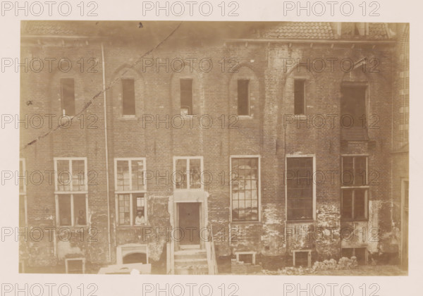 Former St. Agnieten Convent in Utrecht, photograph, annotation: 'Utrecht', annotation: 'Utrecht, Former St. Agnieten Convent now a military building, part bordering a, d church, June 1902', Utrecht, 1902, photographer, Attributed to, paper, pasteboard, albumen print, height 145 mm x width 224 mm, cloisters, monastery, A faded sepia photograph of a brick building facade showing upper and lower rows of windows and doorways, with arched brickwork above the upper windows and rectangular window frames below; the brick surface shows patches of discoloration and weathering, and parts of the plaster or outer layer are chipped away near the base; one narrow vertical pipe runs down the facade; a short exterior stair leads up to a dark doorway in the center with worn steps and a small landing; some windows have multiple panes, some panes are dark or reflective, and a couple of lower windows are open with a person visible at one of them leaning on the sill; the roofline at the top shows tiles and ...