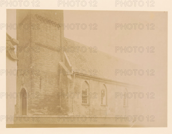 East side of the large church in Vollenhove, photograph, annotation: 'East side of the large church in Vollenhove (Overijsel) Aug 1898', annotation: 'Vollenhove', Vollenhove, 1898, photographer, Attributed to, paper, pasteboard, albumen print, height 171 mm x width 232 mm, church (exterior), A faded sepia-toned photograph of a brick building showing a tall tower corner and a sloped tiled roof, with arched windows set into the wall below the roofline and a narrow doorway at the tower base, a downspout running along the wall, and a short iron fence section visible near the windows.