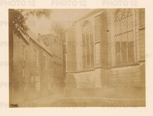 Town hall and the large church in Vollenhove, photograph, annotation: 'Large church and town hall in Vollenhove (Overijsel) Aug 1898', annotation: 'Vollenhove', Vollenhove, 1898, photographer, Attributed to, paper, pasteboard, albumen print, height 167 mm x width 232 mm, church (exterior), townhall, A sepia-toned photograph showing the exterior of adjacent buildings with large arched windows featuring diamond-patterned panes, stone walls with horizontal courses, and brick facades with multiple rectangular windows; a narrow alleyway runs between the buildings, tree branches and leaves appear at the top left, and the foreground shows a paved or stone surface.
