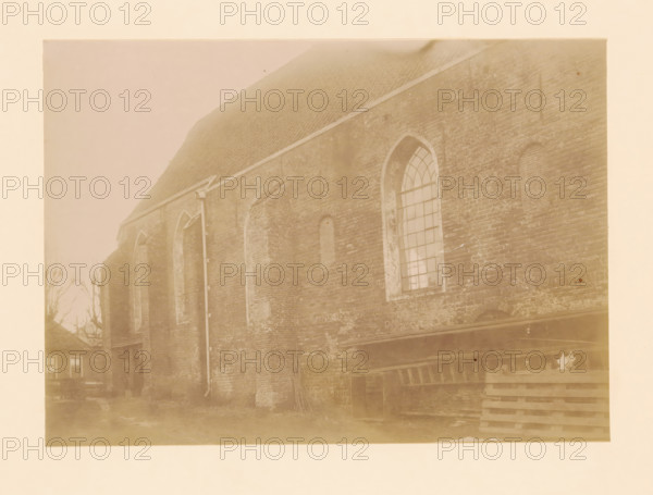 North side of the St. Lambertus Church in Wilsum, photograph, annotation: 'Wilsum', annotation: 'Wilsum North side of the Dutch Reformed Church', Vollenhove, 1897, photographer, Attributed to, paper, pasteboard, albumen print, height 164 mm x width 228 mm, church (exterior), Wilsum, St. Lambertus Church (Wilsum), A faded sepia photograph showing the side of an old brick building with a steep tiled roof and several tall arched window openings, one of which contains a multi-pane window, and others appearing filled or bricked in; a vertical downspout runs along the wall; the brickwork shows weathering and patches of lighter mortar; at the ground level there are stacked wooden pallets and a small open doorway, and in the left background there are bare tree branches and the corner of a smaller building with visible windows.