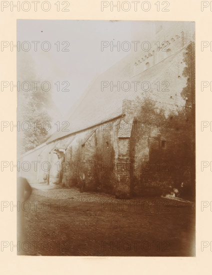 Part of the St. Nicholas Church in Wijhe, photograph, annotation: 'Dutch Reformed Church in Wijhe Aug 1911', annotation: 'Wijhe', Wijhe, 1911, photographer, Attributed to, paper, pasteboard, daylight collodion silver print, height 207 mm x width 156 mm, church (exterior), St. Nicholas Church (Wijhe), A sepia-toned photograph showing a large brick building with a steep tiled roof and decorative brickwork near the top, a cobblestone ground in the foreground, a small arched doorway set into the wall, a projecting roofed entrance with visible wooden supports, a small rectangular window high on the wall, patches of darker discoloration on the bricks, a narrow passage leading left toward a gated area, and tall trees visible faintly behind the building.