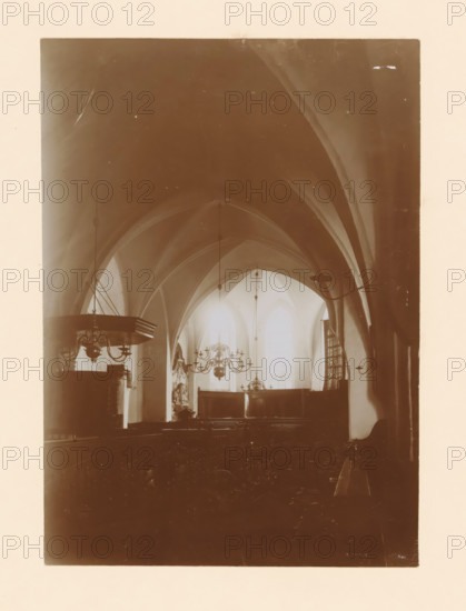 Interior of the St. Nicholas Church in Wijhe, photograph, annotation: 'Wijhe', annotation: 'Dutch Reformed Church in Wijhe Aug 1911', St. Nicholas Church (Wijhe), 1911, photographer, Wijhe, Attributed to, paper, pasteboard, daylight collodion silver print, height 213 mm x width 150 mm, interior of church, A sepia-toned interior scene showing a vaulted ceiling with pointed arches and ribbing, hanging light fixtures and chandeliers suspended from long rods, tall arched windows at the far end allowing light to filter through, dark wooden furnishings including a raised pulpit with ornate detailing on the left and pews extending toward the foreground, decorative wall-mounted lamps on the right, and sculptural or carved elements near the front beneath the windows.