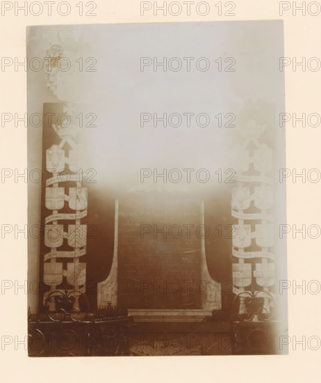 Grave monument of Robert van Ittersum tot Nijenhuis in the St. Nicholas Church in Wijhe, photograph, annotation: 'grafm. van Rober van Ittersum NH kerk Wijhe Aug 1911', annotation: 'Wijhe', St. Nicholas Church (Wijhe), 1911, photographer, Wijhe, Attributed to, paper, pasteboard, daylight collodion silver print, height 208 mm x width 164 mm, Person, Robert van Ittersum tot Nijenhuis, grave-building, monumental tomb, A faded rectangular image with a pale border shows a central dark plaque set within a carved stone frame, flanked on both sides by vertical columns of decorative shields and heraldic motifs; ornate metalwork with curled designs and pointed finials appears along the lower edge, and a pair of carved supports rise beside the plaque; the overall tones are muted sepia with areas of light haze obscuring details toward the top and center.