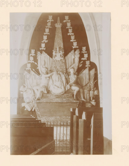 Tomb monument of Coenraad Willem van Dedem in the St. Nicholas Church in Wijhe, photograph, annotation: 'Wijhe', annotation: 'grave monument of Baron van Dedem. Dutch Reformed Church Wijhe Aug 1911', St. Nicholas Church (Wijhe), 1911, photographer, Wijhe, Attributed to, paper, pasteboard, daylight collodion silver print, height 220 mm x width 158 mm, Person, Coenraad Willem van Dedem, grave-building, monumental tomb, A stone memorial set within a tall arched recess features a central tapered obelisk topped with a crown and a carved crest near its upper portion, flanked by draped flags and banners; two seated classical figures, one on each side, hold or touch a large carved shield with a sculpted face and decorative wreath at its center; surrounding the obelisk are multiple small shield-shaped plaques arranged vertically on both sides; the memorial rests on a broad base with molded edges, and the foreground shows rows of wooden pew backs and a metal railing partially obscuring the lower...