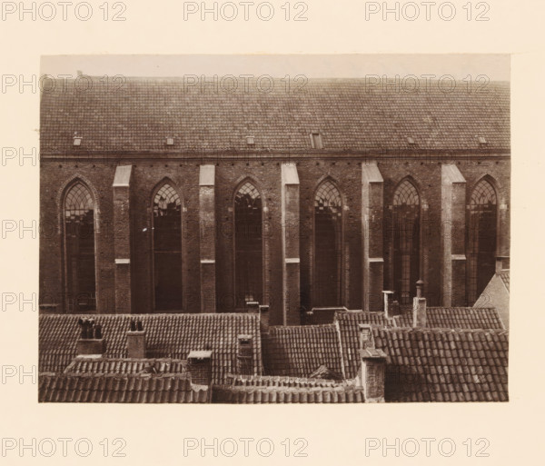 South side of the Broerenkerk in Zwolle, photograph, annotation: 'Broerenkerk, Zwolle', annotation: 'Broerenkerk in Zwolle, South side, June 1909', Zwolle, 1909, photographer, Attributed to, paper, pasteboard, albumen print, height 153 mm x width 208 mm, church (exterior), A sepia-toned photograph showing the upper facade of a large brick building with tall, narrow arched windows set between vertical brick pilasters, a tiled pitched roof above with small rectangular openings, and in the foreground a series of overlapping clay-tiled rooftops with chimneys and vents visible.
