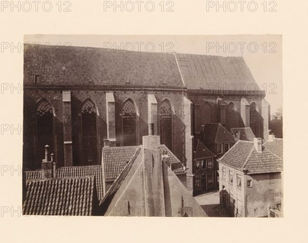 South side of the Broerenkerk in Zwolle, photograph, annotation: 'Broerenkerk in Zwolle, South side, June 1909', annotation: 'Broerenkerk, Zwolle', Zwolle, 1909, photographer, Attributed to, paper, pasteboard, albumen print, height 151 mm x width 218 mm, church (exterior), A sepia-toned photograph showing the upper portions of several buildings clustered together, dominated by a large brick structure with tall arched windows and a steep tiled roof, nearby smaller buildings with steep, tiled roofs and chimneys, visible rooflines and ridges creating angled shapes, textured roof tiles and brick walls, vertical downpipes and buttresses on the large building, shadowed window openings and panes in some of the facades, and a pale blank border surrounding the image.