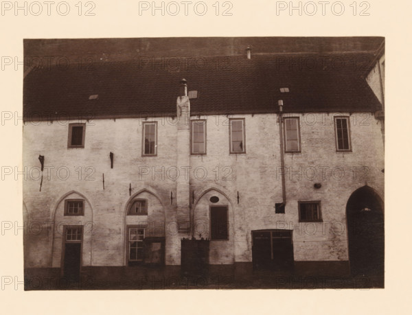 Monastery near the Broerenkerk in Zwolle, photograph, annotation: 'Broerenkerk in Zwolle, cloister north of the church in the barracks June 1909', annotation: 'Broerenkerk, Zwolle', Zwolle, 1909, photographer, Attributed to, paper, pasteboard, albumen print, height 155 mm x width 221 mm, cloisters, monastery, Broerenkerk, A faded, sepia-toned photograph of a brick building facade with a steep tiled roof, small roof vents, and a central chimney running down the wall; the upper wall has tall, narrow windows with muntins, while the lower wall features arched recesses, rectangular windows, and doorways of varying sizes; vertical downspouts and metal fixtures are attached to the exterior, and there is a dark arched passage visible at one side.