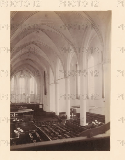 Interior of the Broerenkerk in Zwolle, photograph, annotation: 'Broerenkerk in Zwolle, Interior June 1909, viewed towards the choir', annotation: 'Broerenkerk, Zwolle', Broerenkerk, 1909, photographer, Zwolle, Attributed to, paper, pasteboard, albumen print, height 220 mm x width 157 mm, interior of church, A high vaulted interior with pointed arches and ribbed ceilings supported by tall smooth columns, large arched windows along the side letting in diffuse light, rows of wooden pews and chairs arranged in a central floor area, low wooden partitions creating aisles and enclosures, several multi-globe light fixtures mounted on stands, a raised area at the far end with larger windows behind it, and ornamental capitals where arches meet the columns.