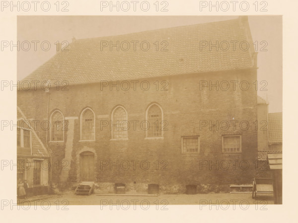 View of the synagogue in Zwolle, photograph, annotation: 'Synagoge, Zwolle', annotation: 'Zwolle synagogue March 1899 (O.IJ. is being demolished', Zwolle, 1899, photographer, Attributed to, paper, pasteboard, albumen print, height 166 mm x width 233 mm, the Synagogue (as building and place of worship, Jewish religion), A sepia-toned photograph of a large brick building with a steep tiled roof and several arched windows set into the upper wall, rectangular windows lower down, and a tall wooden door with a small stepped platform in front; a narrow vertical downpipe runs along the facade, and low arched openings sit at the base of the wall; to the left a smaller gabled structure with visible windows and a woman standing near its entrance, and to the right a partial view of another tiled roof and a wooden bench or cart against the wall.
