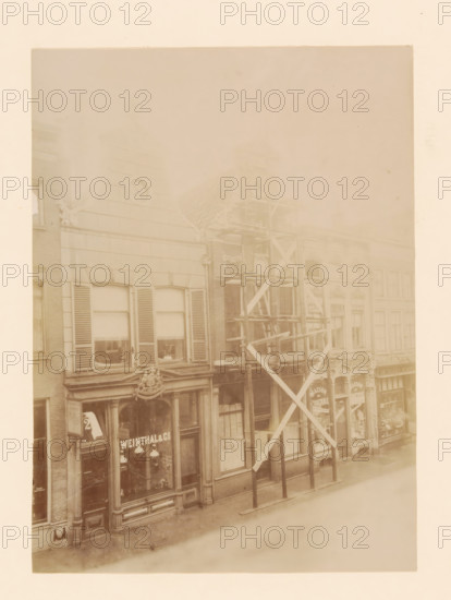 Diezerstraat 43 in Zwolle, photograph, annotation: 'Zwolle', annotation: 'Woonuis Diezerstraat 43 Zwolle (O.IJ.) Dec: 1905', Zwolle, 1905, photographer, Attributed to, paper, pasteboard, albumen print, height 230 mm x width 168 mm, urban housing, street, A faded sepia photograph of a row of multi-story storefront buildings with large windows and decorative architectural details, one facade showing shuttered upper windows and a triangular pediment with an emblem, another facade partially covered by wooden scaffolding with diagonal supports and vertical posts, carved ornamental moldings above the ground-level shop windows, display windows revealing hanging lamps and interior shelving, narrow sidewalks in front of the buildings, and a pale sky above.