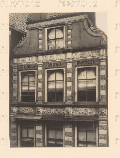 Façade of Sassenstraat 33 in Zwolle, photograph, annotation: 'Zwolle', annotation: 'Zwolle. House Sassenstraat 33. detail. Oct 1907', Zwolle, 1907, photographer, Attributed to, paper, pasteboard, gelatin silver print, developed gelatin silver print, height 220 mm x width 161 mm, urban housing, façade (of house or building), A sepia-toned photograph of a building facade showing ornate architectural details with three tall arched windows framed by decorative pilasters, patterned stonework on the pillars, carved floral and foliate friezes between the floors, and sculpted faces and reliefs integrated into the ornamentation; the upper section includes a rounded window with patterned curtains and a curved pediment flanked by reclining sculpted figures, while the lower sections show window panes with blinds or curtains and a projecting awning above the ground-floor opening.