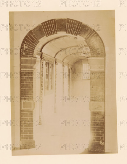 Covered hall in the Women's House in Zwolle, photograph, annotation: 'Zwolle hallway in the women's house on the Melkmarkt Sept 1893', annotation: 'Zwolle', Zwolle, 1893, photographer, Attributed to, paper, pasteboard, albumen print, height 232 mm x width 171 mm, vestibule, hall, old-people's home, A sepia-toned photograph of an arched corridor framed by brick and stone columns, showing a curved vaulted ceiling with decorative moldings and carved rosettes, a section of a window with small panes at the far end, ornate pilasters and carved vertical embellishments along the walls, and a floor and lower walls that fade toward the foreground.