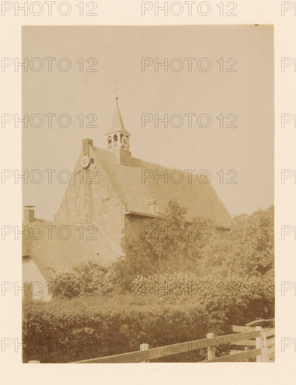 View of the church of Windesheim, photograph, annotation: 'Windesheim (O.IJ.) Church of the Dutch Reformed congregation May 1901', annotation: 'Windesheim, municipality of Zwollerkerspel', Windesheim, 1901, photographer, Attributed to, paper, pasteboard, albumen print, height 226 mm x width 169 mm, church (exterior), A sepia-toned photograph showing the upper portion of a building with a steep tiled roof and a small belfry topped by a pointed spire and a weather vane, the brick gable end featuring a circular decorative element and a chimney, a dormer window set into the roof, dense trees and shrubs in front of the building, and a wooden fence in the foreground.