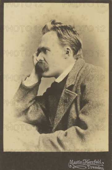Portrait of Friedrich Nietzsche, photograph, annotation: 'Nietzsche', photographer: Martin Herzfeld, Dresden, c. 1882, 1910, Saxony, paper, pasteboard, daylight gelatin silver print, height 143 mm x width 101 mm, height 168 mm x width 109 mm, Person, Friedrich Nietzsche, historical persons, A sepia-toned portrait of a man shown in profile looking to the left, with a high forehead and swept-back hair, a thick mustache, and a hand resting against his cheek; he wears a collared shirt, a dark tie or vest beneath, and a textured jacket with a visible lapel and pocket, all set against a plain, lightly speckled background with a dark border around the image.
