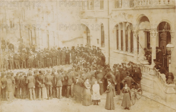 Soldiers in front of the town hall of Valkenburg, Headquarters maneuvers in Limburg 1901, done in Valkenberg, cabinet photograph, collector's mark: 'Vosmaer', stamp: 'Photographic Atelier, von, Joh. Cohnen, Valkenburg, Groote Straat 130', photographer: Johan Joachim Cohnen, Valkenburg (Limburg), 1901, Johan Joachim Cohnen, photographer, paper, pasteboard, baryta paper, daylight collodion silver print, height 106 mm x width 167 mm, maneuver, military training, military parade, pageant, A large sepia-toned street scene shows a dense crowd of people gathered outside a stone building with arched windows and decorative columns; many men in uniform and hats stand clustered together, some in lines and some facing toward the building, while a few women in long dresses and high collars stand nearer the foreground, one holding a bicycle with a large front wheel. The building features a balcony area where several figures lean on the balustrade and others stand near an ornate lamp post, and a grou...