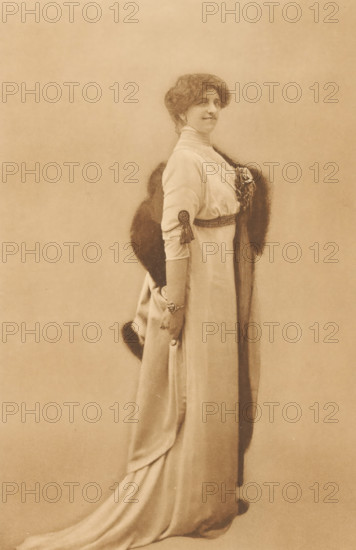 Portrait of Anna de Savornin Lohman, photomechanical print, annotation: 'Mevrouw, A. de Savornin -, - Lohman', name: 'Foto Ebner, The Hague.', photographer: Ferdinand Robert Ebner, The Hague, cliché maker: anonymous, in or after 1890, paper, collotype, photoprint, height 200 mm x width 130 mm, Person, Anna de Savornin Lohman, portrait of a writer, A sepia-toned portrait of a woman standing in profile, wearing a long, flowing gown with a high collar and fitted sleeves, a dark fur stole draped over her shoulders, a decorative belt at her waist, a tassel ornament on her sleeve, a bracelet on her wrist, and her hair styled in a voluminous updo while she looks slightly toward the camera with a faint smile.