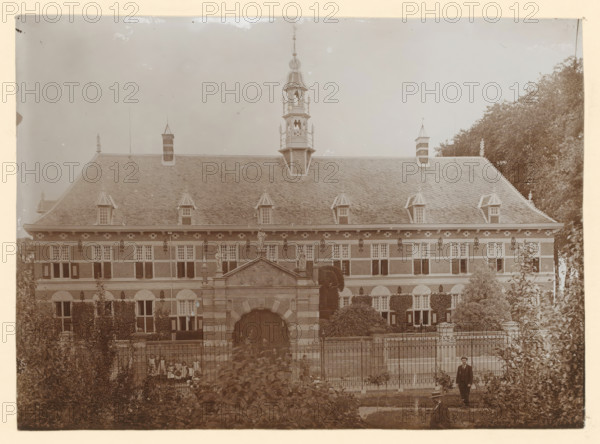 View of the Royal Orphanage in Buren, photograph, annotation: 'Orphanage, Buren', annotation: 'The Orphanage in Buren founded by deed of endowment, ', Buren, 1890 - 1920, photographer, paper, pasteboard, daylight collodion silver print, height 424 mm x width 300 mm, orphanage, Royal Orphanage, A large multi-story building with a steep tiled roof and multiple dormer windows dominates the image, topped by a decorative tower with an ornate spire; the façade shows regularly spaced windows with shutters and decorative stonework, and a central arched entrance framed by sculptural elements and a pediment; in front of the building there is a wrought iron fence and a formal garden area with hedges and trees, and a man in a dark suit stands near the lower right foreground.