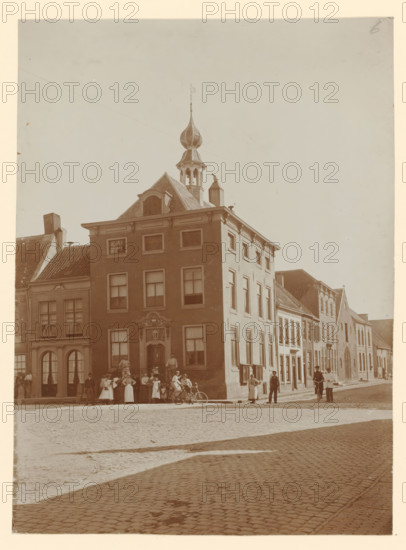 View of the town hall in Buren, photograph, annotation: 'Stadhuis, Buren', annotation: 'Het stadhuis te Buren', Buren, 1890 - 1910, photographer, paper, pasteboard, daylight collodion silver print, height 424 mm x width 295 mm, townhall, A tall brick building with a decorative entrance and multiple windows stands on a cobblestone street, topped by a roof structure with a small tower and an onion-shaped spire, flanked by neighboring buildings of varying heights and roof styles; groups of people, some standing and some seated on a bench or bicycles, gather on the sidewalk in front of the buildings, while a few individuals are seen walking or standing along the street, with shadows cast across the cobbles and an open sky above.