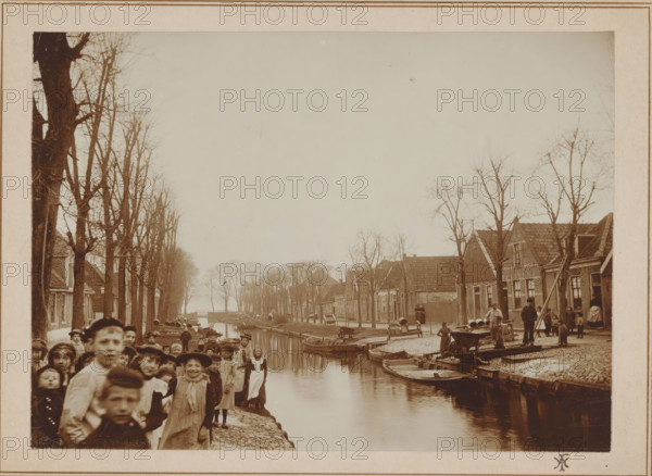 View of a canal in Edam, photograph, annotation: 'Vaart te Edam', monogram: 'HI', photographer: unknown, Edam, 1880 - 1910, paper, pasteboard, daylight collodion silver print, height 159 mm x width 208 mm, canals, waters (in village), street (city(-scape) with figures, staffage), A sepia-toned photograph of a canal scene lined with leafless trees and rows of houses on both sides, with small boats moored along the canal edges and people standing on the banks and near the water; a group of children in the foreground look toward the camera, some wearing hats and simple clothing, while adults and other children are scattered along the right-hand bank where wooden planks, barrels, and carts are visible, and a narrow bridge spans the canal in the distance under a pale sky.