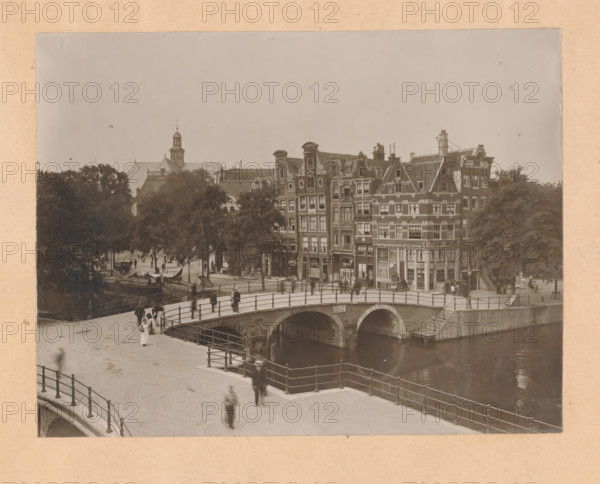 View of the Prinsengracht at the Brouwersgracht in Amsterdam, photograph, annotation: 'Prinsengracht, Brouwersgr.', annotation: 'Houses purchased by the association, Hendrik de Keyser, Prinsengracht b, d Brouwersgracht, photo ...', number: ''19.10', Amsterdam, 1900 - 1920, photographer, paper, pasteboard, gelatin silver print, developed gelatin silver print, height 244 mm x width 320 mm, urban housing, village, canals, waters (in city) (city(-scape) with figures, staffage), Prinsengracht, A sepia-toned scene shows a curved stone bridge with iron railings spanning a calm canal beneath brick arches, pedestrians walking along the cobbled street and across the bridge, blurred figures indicating motion, a row of tall narrow multi-story buildings with decorative gables and many windows lining the canal, small shopfronts at street level, leafy trees behind and between the buildings, boats moored along the canal near the trees, and a distant church tower rising above the rooftops.