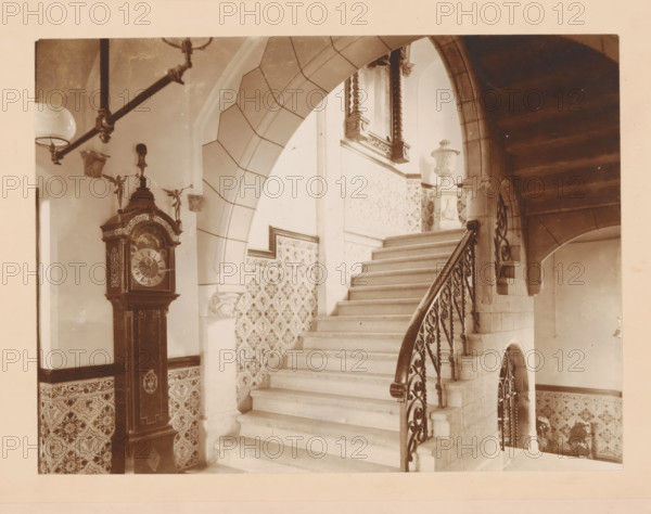 Staircase in the former Groninger Museum, photograph, annotation: 'VI, ZH, Museum Groningen', number: '1380', Groningen (city), 1880 - 1910, photographer, paper, pasteboard, daylight collodion silver print, height 299 mm x width 358 mm, staircase, A wide stone staircase with a curved wrought-iron handrail rises through an arched opening, flanked by patterned tiled walls and a stone newel supporting an ornate carved urn, while a tall grandfather clock with decorative figures and carved details stands against a tiled lower wall beneath a hanging light fixture and a ceiling beam.