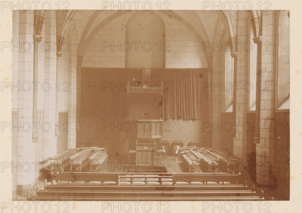 Interior of a hall at the University of Utrecht, photograph, inscription: 'AW van Leeuwen. Utrecht', retailer's mark: 'G. Hilhorst A zn, Acad boek en kunsthandel, Algem. Muziekh., Domsteeg F.1, Utrecht', annotation: 'int. Universiteit Utrecht', number: '09' 79', photographer: AW van Leeuwen, Utrecht, 1890 - 1909, paper, pasteboard, daylight collodion silver print, height 228 mm x width 305 mm, university, Universiteit Utrecht, A sepia-toned interior view of a vaulted room with tiled walls and pointed arches supported by tall columns with decorative capitals, rows of wooden chairs with woven seats arranged facing a central wooden lectern or pulpit, a raised area at the back with a curtained backdrop and a decorative wooden canopy above the pulpit, narrow tall windows set between the columns, patterned lower wall panels, and scattered small objects and wear marks visible on surfaces.