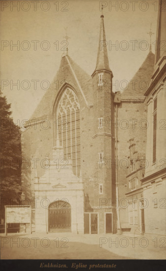 Part of the Zuiderkerk in Enkhuizen, photograph, inscription: 'Enkhuizen, Eglise protestante', Enkhuizen, 1870 - 1900, photographer, paper, pasteboard, albumen print, albumendruk, height 195 mm x width 123 mm, church (exterior), A sepia-toned photograph of a large brick church facade with a tall pointed spire and steep gabled roof, featuring a large pointed arched stained-glass window with tracery set into the wall, a square brick tower with narrow slit windows rising beside the window, and an ornate stone entrance with a gated opening at ground level; adjacent buildings with windows and decorative masonry are visible on the right, a tree tops appear on the left, and a paved area lies in front of the structures.