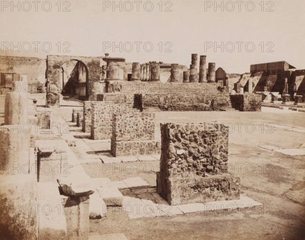 Tempio di Giove, Ruin of the Temple of Jupiter at Pompeii, Pompei, photograph, collector's mark: 'VOSMAER', number: '1208.', photographer: Giorgio Sommer, Pompeii, 1863 - 1875, Giorgio Sommer, photographer, pasteboard, paper, albumen print, height 314 mm x width 420 mm, ruin of a building, architecture, landscape with ruins, temple, shrine, non-Christian religions, Temple of Jupiter (Pompeii), A sepia-toned photograph of an archaeological site showing rows of stone structures and foundations on a flat open area, with rough rectangular stone blocks in the foreground and a pathway of stone slabs leading toward a raised platform; on the platform there are several fluted stone columns, some standing and some partially intact, and a large brick archway is visible to the left of the platform; in the background there are low stone walls and additional ruins with stair-like terraces and scattered stone fragments, all under a pale sky.